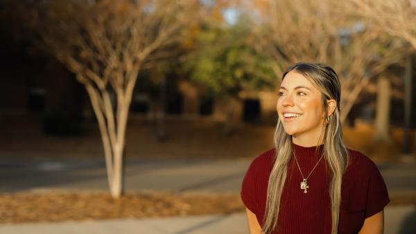 Women wearing maroon top looking to her right standing in front of trees.